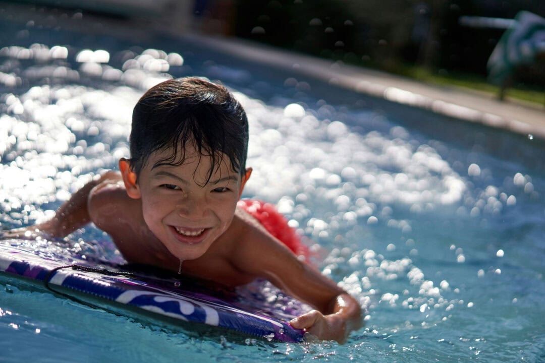 Young boy wearing red swim trunks lying on his stomach on a blue paddle in an inground swimming pool. Behind the boy the water reflects the sunlight.