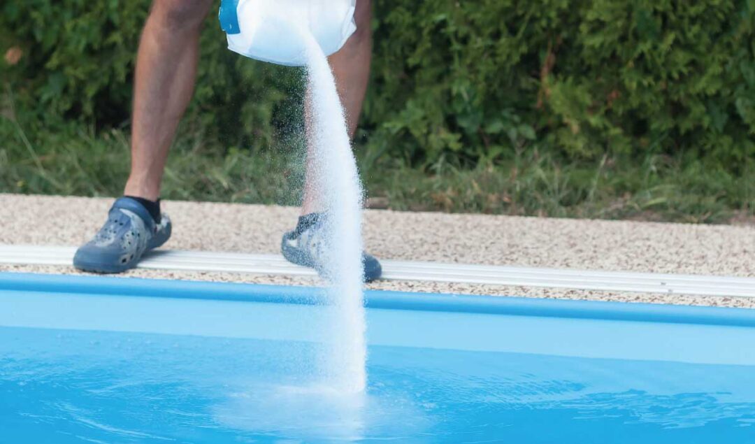 A medium shot of salt being poured from a bag into an inground pool. The pool makes up the bottom third of the image. Legs can be seen in front of a wooded area directly behind the salt.