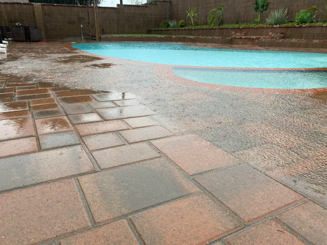A low-angle wide shot of an inground pool. The pool water is overflowing onto the already wet concrete due to excessive heavy rain.