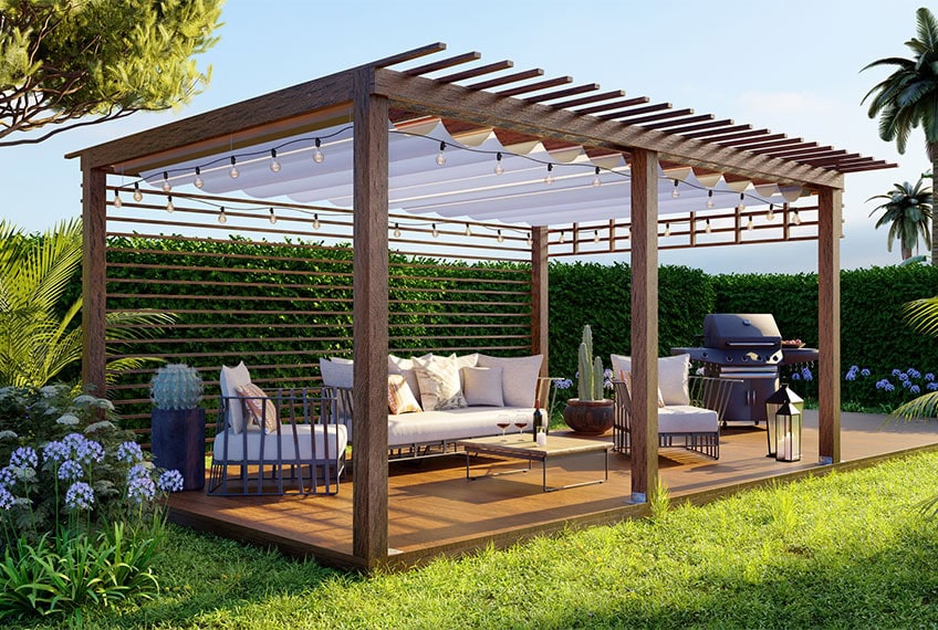 A pergola shade structure with a tarp hanging under the ceiling. under the pergola are two chairs, a loveseat, and a grill. The pergola sis surrounded by purple plants and a hedge behind it. 