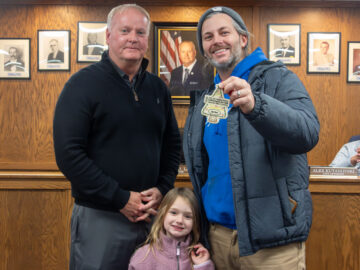 Two men pose for a picture in front of a wood wall with multiple pictures while a young girl stands between them. The man on the right holds a tree-shaped plaque. All three are smiling. Another man can be seen sitting to the right of them cut off by the edge of the image.