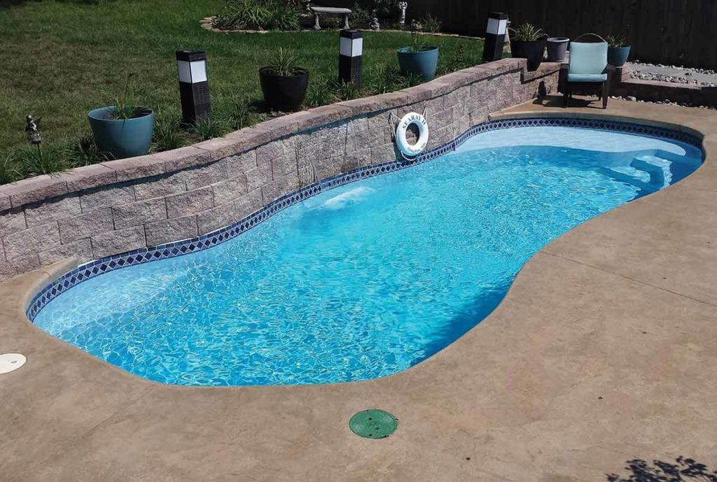A light-blue freeform fiberglass pool featuring a retaining wall above the backside wall. The retaining wall is spilling water into the pool from the top in the middle. The front and sides of the pool is surrounded by concrete. Grass and potted plants are above the retaining wall in the grass.