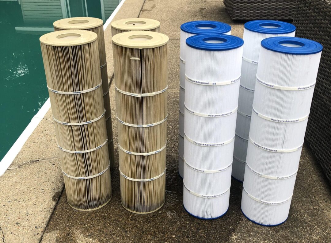 Eight pool cartridge filters side-by-side next to a pool. The four cartridges closest to the pool on the left are stained yellow and brown. The four cartridges on the right are bright white. 